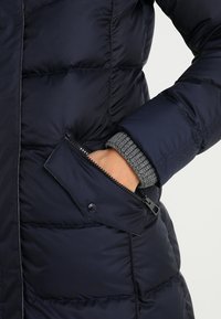 Marinblå puffjacka med kviltad struktur och blixtlåsficka på sidan. Har en grå stickad manschett synlig vid handleden.