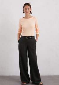 Helles Pfirsichfarbenes Langarmshirt mit weitem Ausschnitt, kombiniert mit schwarzen Weitbein-Hosen und einem schwarzen Gürtel mit goldener Schnalle.