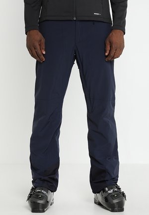 Marineblauwe waterdichte skibroek met ritssluitingen in de zakken, elastische manchetten en duurzame stof; gecombineerd met zwarte skischoenen met verstelbare gespen.