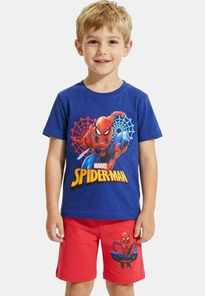 Garçon blond souriant, portant un t-shirt bleu Spider-Man avec des motifs de toile d'araignée et un short rouge affichant un motif de Spider-Man sur la jambe gauche.