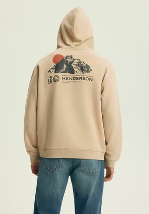 Osoba nosząca beżową bluzę z kapturem z grafiką góry, czerwonym słońcem i napisem „HENDERSON” na plecach, w połączeniu z niebieskimi dżinsami.