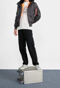 Pánská bomber bunda tmavě šedé barvy s oranžovou podšívkou, doplněná světlejší tričkem s potiskem, černými kalhotami a šedými teniskami. Stojí na stříbrné krabici.