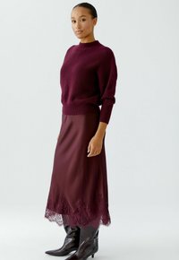 Burgunderfarbener Strickpullover mit hohem Kragen, kombiniert mit einem seidig glatten burgunderfarbenen Rock mit Spitzenbesatz; getragen mit glänzenden schwarzen kniehohen Stiefeln.