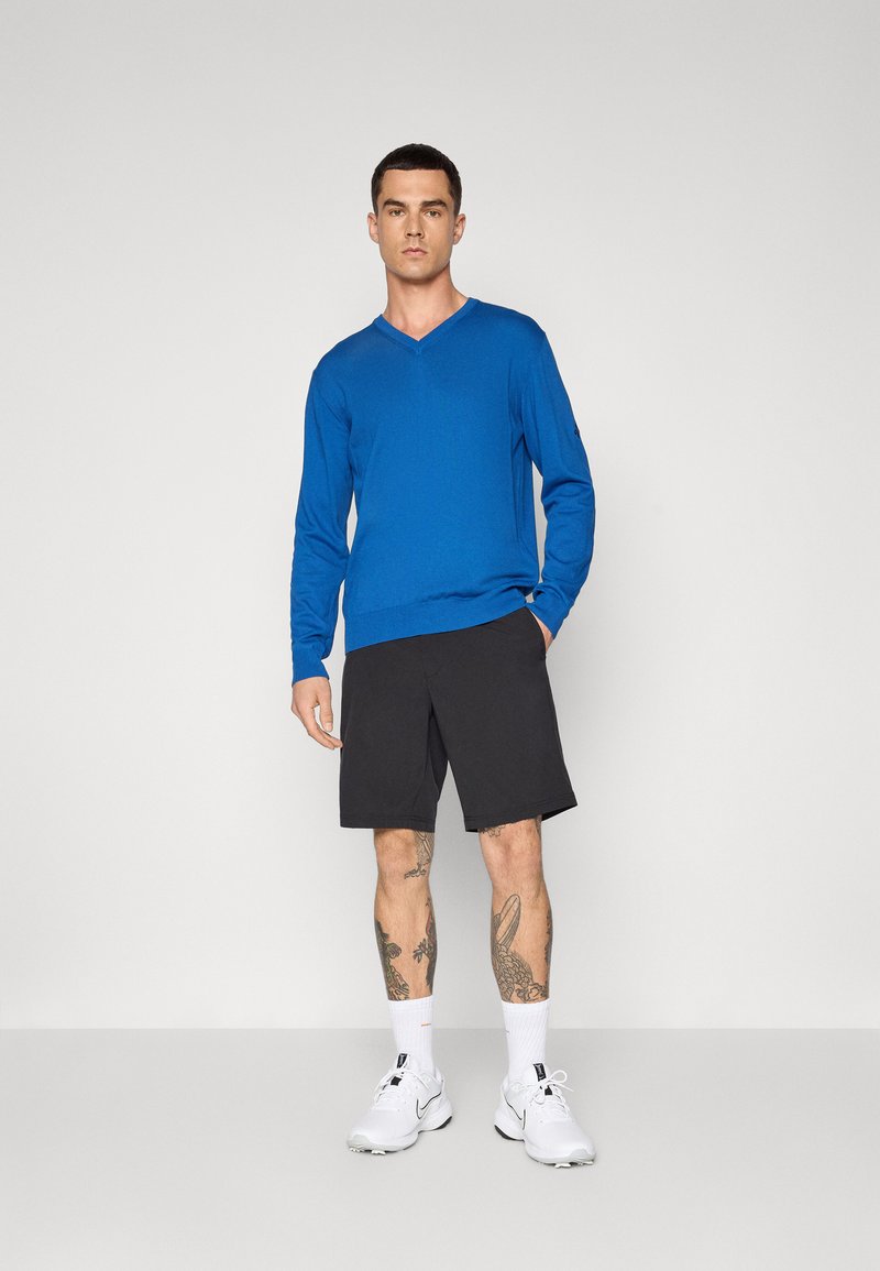Blauwe v-halstrui, lichte brei, gecombineerd met zwarte shorts en witte sportschoenen. Zichtbare tatoeages op de onderbenen. Neutrale achtergrond.