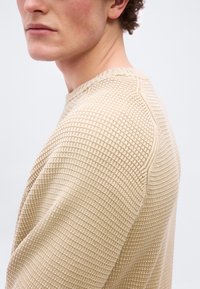 Homme aux cheveux bouclés portant un pull en tricot texturé beige, vu de profil sur un fond neutre.