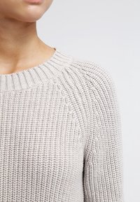 Nahaufnahme einer Person, die einen hellgrauen, grob gestrickten Pullover mit einem gerippten Rundhalsausschnitt vor einem schlichten Hintergrund trägt.