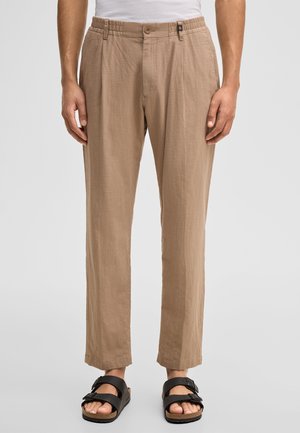 Beige gerade geschnittene Hose mit Knopf und elastischem Bund, getragen mit schwarzen Sandalen mit Schnallen und einem weißen Oberteil, gezeigt von der Taille bis zu den Füßen.