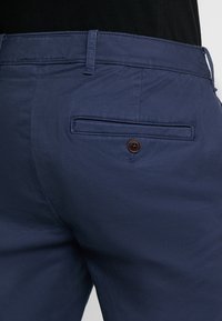 I pantaloni blu navy presentano una texture liscia, una vita standard con passanti per cintura e una tasca posteriore adornata con un bottone marrone.