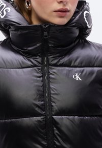 Μαύρο μπουφάν τύπου puffer με ψηλό γιακά, κλείσιμο με φερμουάρ και γυαλιστερό φινίρισμα. Διαθέτει διακοσμητικό λογότυπο στο στήθος και λεία υφή.