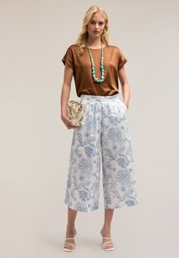 Top corto in raso marrone con maniche corte abbinato a culotte bianche con motivi floreali blu. Gli accessori includono una collana di perle blu e una borsa crema.