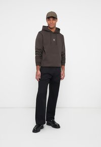 Homme portant un hoodie marron avec un symbole des Gémeaux, un pantalon noir, des chaussures noires et une casquette à carreaux, se tenant devant un fond blanc.