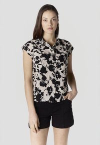 Mladá žena s dlhými tmavými vlasmi, oblečená v čiernobielej kvetinovej blúzke s krátkymi rukávmi a čiernych krátkych nohaviciach, stojaca s jednou rukou v vrecku.