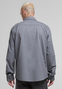 Szürke gombos ing gallérral, hosszú ujjakkal és gombos mandzsettákkal. Az anyag simának tűnik, matt textúrájú, minták nélkül.