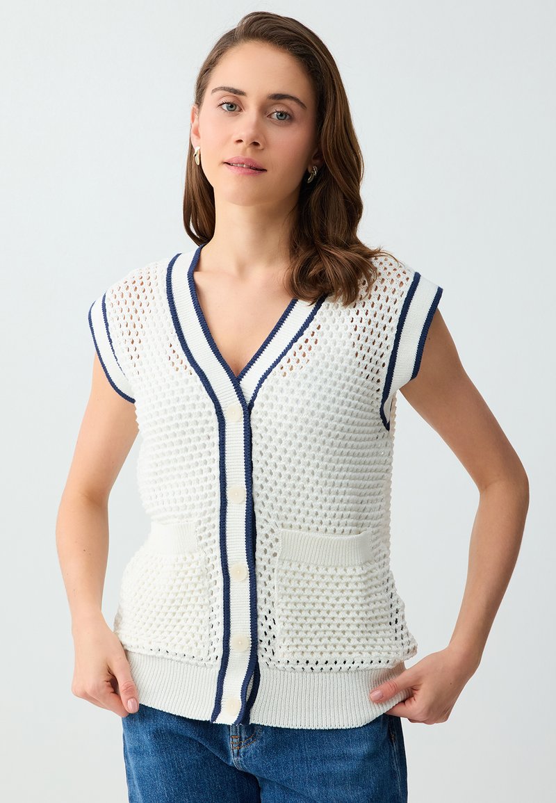 Femme portant un gilet tricoté sans manches blanc avec bordure et boutons bleu marine, associé à un jean bleu, debout devant un fond uni.