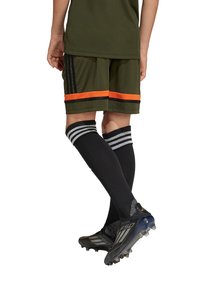 Schwarze Fußballschuhe, olivgrünes Trikot und Shorts mit orangefarbenen und schwarzen Streifen, schwarze knielange Socken mit weißen Streifen, strukturiertes Material.