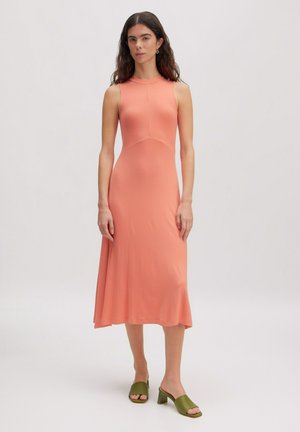 Femme debout portant une robe midi corail sans manches avec des sandales vertes à talon bloc ouvert à l’orteil devant un fond clair uni.