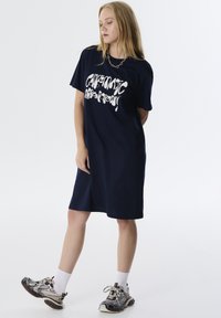 Marineblauw oversized t-shirt jurk met witte grafische tekst. Korte mouwen, rechte snit, zachte katoenstof en een relaxte pasvorm.