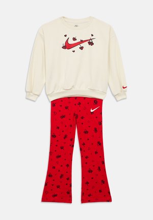 Bluza crem cu logo Nike Swoosh roșu și grafică cu cireșe și inimi, asortată cu pantaloni roșii cu modele negre de cireșe și inimi și logo alb Nike.