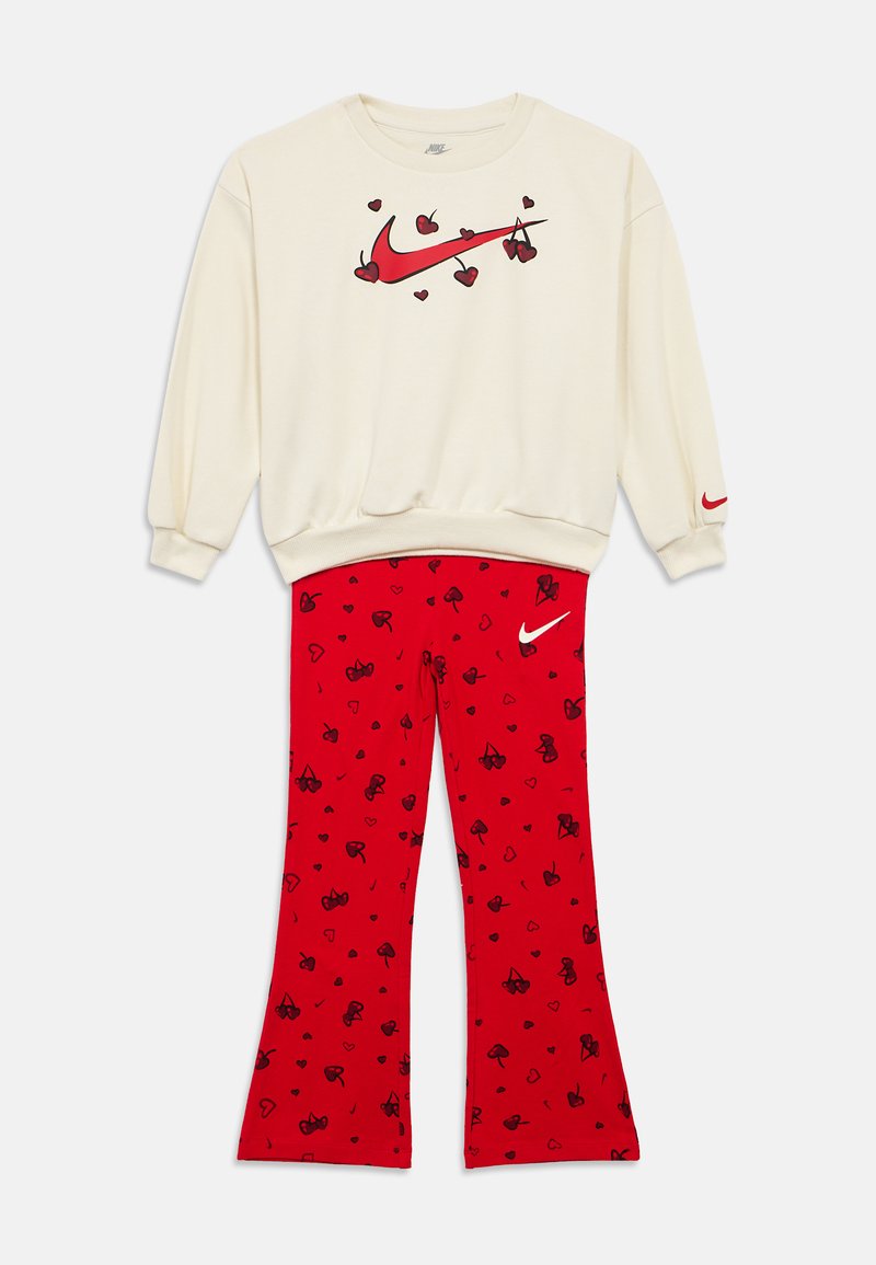 Φούτερ εκρού με κόκκινο logo Nike swoosh και γραφικά με κερασάκια σε σχήμα καρδιάς, συνδυασμένο με κόκκινο παντελόνι που έχει μαύρα κεράσια και σχέδια καρδιάς καθώς και λευκό λογότυπο Nike.