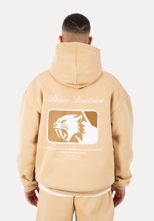 Man wearing beige hoodie and sweatpants, back view, hoodie features white graphic of roaring panther and text "Athleisure Department."