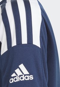 Marineblå sportsjakke med tre lodrette hvide striber på skuldrene, mønstret stof og et hvidt Adidas-logo på brystet.