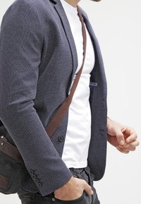 Marinblå texturerad kavaj med slim fit. Bärs över en vit t-shirt, tillsammans med mörka jeans. Brun crossbody-väska syns på vänster sida.