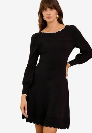 Black knit dress with long sleeves, scalloped neckline, and a flared skirt. Features textured patterns on sleeves and hem.
