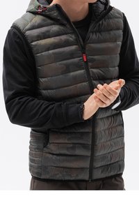 Gepolstertes Weste mit Kapuze, schwarzes und dunkelgrünes Camouflage-Muster, Reißverschluss und rote Akzente. Über einem langärmligen schwarzen Shirt getragen.