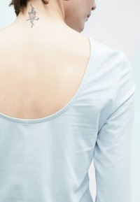 Ανοιχτό μπλε τοπ με μακριά μανίκια και ευρύ ντεκολτέ στην πλάτη. Μαλακό βαμβακερό ύφασμα με λεία υφή. Διακρίνεται εμφανώς ένα τατουάζ στον λαιμό.