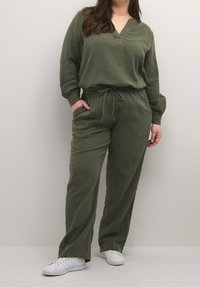 Olivengrünes Zweiteiler-Outfit aus strukturiertem Material. Mit einem V-Ausschnitt-Top, elastischem Bund und weitem Bein. Kombiniert mit weißen Sneakers.