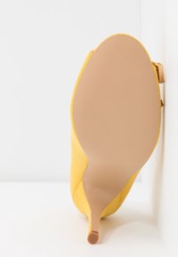 Chaussure à talon haut jaune avec un dessus en tissu texturé et un bout pointu. La semelle est élégante et lisse, affichant une couleur beige.