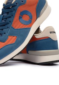 Chaussures de sport bleues et orange avec semelles blanches épaisses, lacets bleu marine et logo circulaire sur le côté, montrant des vues latérales et arrière.