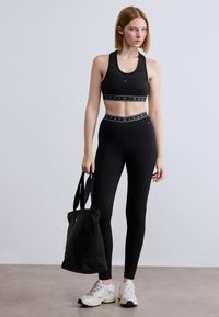 Μαύρο αθλητικό crop top με λωρίδα λογότυπου, ταιριαστά κολάν με ψηλή μέση και μαύρη τσάντα tote. Το μοντέλο φορεί άσπρα αθλητικά παπούτσια.