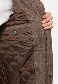 Braune Steppjacke mit Fischgrätmuster außen und glattem Innenfutter, mit einem Etikett, das in der Innentasche sichtbar ist.