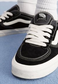 Black suede sneakers with white accents, featuring white laces, stitching details, and a round toe, set against a blue background.