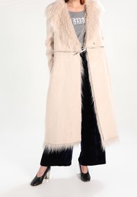Długi kremowy płaszcz z syntetycznym futrem przy kołnierzu i u dołu, z wiązaniem w talii i miękką fakturą, połączony z ciemnymi aksamitnymi spodniami i czarnymi szpilkami.