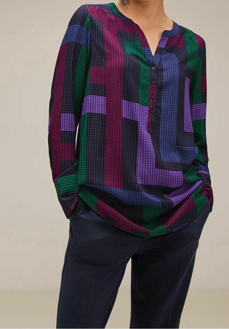 Personne portant un chemisier à manches longues avec des motifs géométriques verts, violets, bleus et noirs, rentré dans un pantalon foncé, les mains dans les poches.