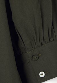 Poignet en tissu vert foncé avec plis froncés et deux boutons, un noir et un blanc, cousus près du bord.