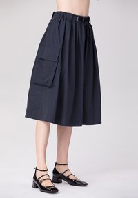 Jupe mi-longue noire en tissu léger, avec une taille ceinturée, un bandeau élastique et une poche latérale. Associée à des chaussures noires en vernis.