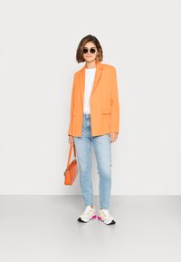 Blazer ajustado laranja sobre uma t-shirt branca, jeans azul claro e ténis robustos. Os acessórios incluem uma pequena mala laranja e óculos de sol escuros.