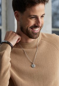 Pull en beige à côtes, collier en chaîne argentée avec un pendentif circulaire, et un bracelet en cuir marron porté au poignet.