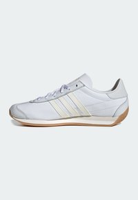 Vit sportskor i läder med texturerade detaljer, med tre diagonala ränder. Ljust grå sula med en platt design och minimalistisk snörning.