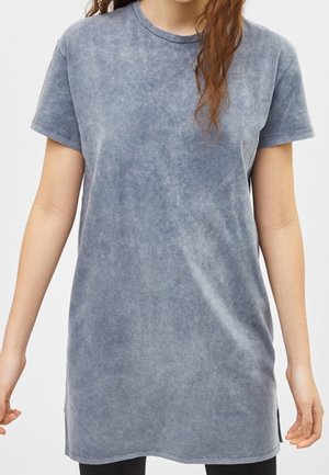 Kobieta ubrana w wyblakły niebieski, za duży t-shirt z krótkim rękawem, stojąca z ramionami swobodnie opuszczonymi wzdłuż ciała.