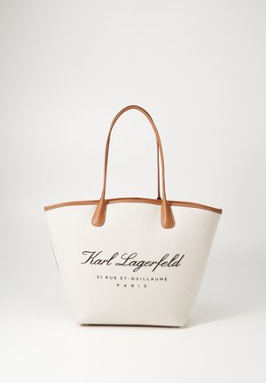 Canvas tote bag with brown leather accents. Features logo and text in dark brown on a cream background. Rounded shape with long handles.