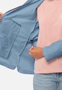 Lichtblauwe jas met een fijne textuur, voorzien van een binnenzak en een verstelbaar trekkoord; gedragen over een roze T-shirt en denim jeans.