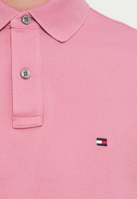 Rožinė polo marškinėlių, pagaminti iš tekstūruotos medžiagos, su klasikinio kirpimo apykakle, dviem mygtukais ir mažu išsiuvinėtu logotipu kontrastingomis spalvomis.