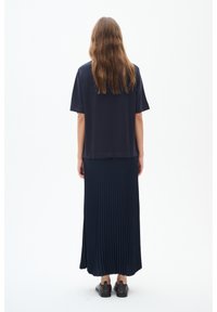 Granatowa bluzka z krótkim rękawem w połączeniu z długą, plisowaną spódnicą, obie wykonane z miękkiego materiału. Zawiera boczne rozcięcia i gładką fakturę. Czarne buty.