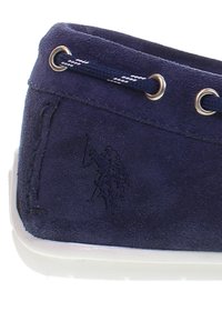 Mörkblå mockaskor med vit sula, med metallöglor och mörkblå snören, präglad logotyp av en ryttare på häst.