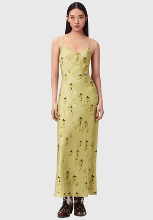 Kvinde med langt mørkt hår iført en gul satin natkjole med blomsterprint og brune og hvide støvler i ko-skind, stående mod en ensfarvet baggrund.