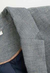 Tkaninowa kurtka o fakturze w deseń houndstooth w granatowo-białych kolorach. Posiada beżową podszewkę. Zbliżenie na kołnierz i rękaw.